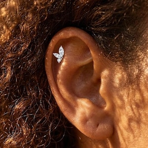May include: A close-up of an ear featuring a butterfly-shaped earring. The earring is made of small, sparkling stones, possibly diamonds or cubic zirconia, set in a silver or white gold setting. The earring is placed on the upper ear.