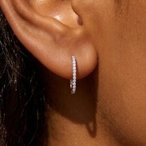 May include: A close-up of a small, silver hoop earring. The earring is set with a row of small, clear gemstones. The earring is in the ear of a person with brown skin.