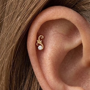 May include: A gold ear stud featuring a curved, leaf-like filigree design that extends upward from a single, round, clear gemstone. The jewelry is positioned in the upper cartilage area of the ear, showcasing a polished metallic finish.