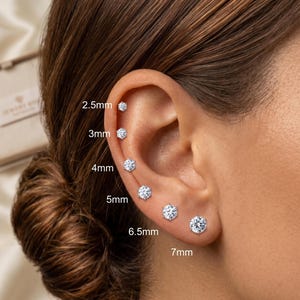 May include: A set of six silver-toned stud earrings with clear round stones, arranged in ascending order of size along the earlobe and cartilage. The sizes are labeled as 2.5mm, 3mm, 4mm, 5mm, 6.5mm, and 7mm, showing the diameter of each individual earring.