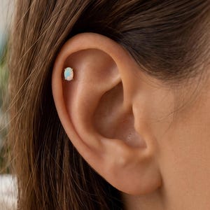 May include: An oval-shaped opal stud earring with a gold-toned setting, positioned in the upper cartilage of the ear. The gemstone displays iridescent flashes of white, blue, and pink light against a smooth, polished surface in a prong setting.