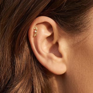 Pode incluir: Um brinco de cartilagem em tom dourado com um design de filigrana curvo e intrincado, apresentando uma pequena pedra preciosa transparente de corte redondo cravada perto do topo. A peça está posicionada na cartilagem superior da orelha.