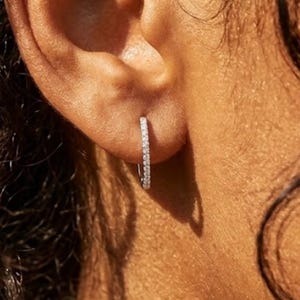 May include: Close-up of a small, silver hoop earring. The earring is encrusted with small, clear gemstones. The earring is in the ear of a person with brown skin. The earring is a classic jewelry piece.