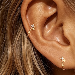 Peut inclure: Deux boucles d'oreilles dorées présentant un motif en forme d'étoile avec une petite pierre claire au centre de l'étoile supérieure. Chaque boucle se compose de deux étoiles reliées par une fine chaîne, créant un effet pendant sur l'oreille.