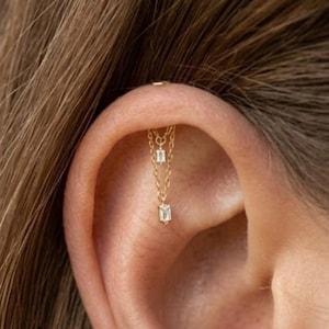 Könnte beinhalten: Ein goldener Kettenohrring mit zwei zarten, hängenden Ketten, die an einem einzigen Stecker befestigt sind. Jede Kette ist mit einem kleinen, klaren, rechteckigen Edelstein im Baguetteschliff verziert. Der Schmuck sitzt im oberen Knorpelbereich.