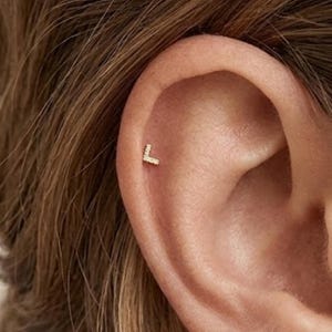 May include: A small gold-toned stud earring shaped like the capital letter L, featuring a pavé-set surface with tiny clear stones. The earring is positioned in the upper cartilage area of the ear, displaying a metallic finish and a geometric design.