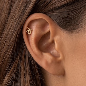 May include: A gold-toned metal earring shaped like a small, coiled snake with a textured surface. The earring is positioned in the upper cartilage area of the ear, featuring a curved body and a distinct head, secured with a small post backing.