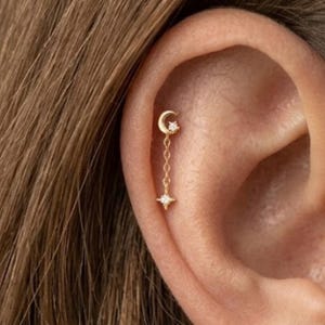 May include: A gold cartilage earring featuring a crescent moon stud with a small clear stone, connected by a delicate chain to a dangling four-pointed star charm set with a central clear stone. The jewelry is positioned on the upper ear cartilage.