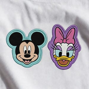 Peut inclure: T-shirt blanc avec des écussons brodés de Mickey Mouse et Daisy Duck. Mickey a un visage noir avec un contour bleu clair. Daisy Duck a un contour et un nœud violets.