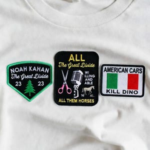 Puede incluir: Camiseta blanca con tres parches bordados. Los parches muestran texto y gráficos: "NOAH KAHAN The Great Divide 23", "ALL The Great Divide ALL THEM HORSES", y "AMERICAN CARS KILL DINO".