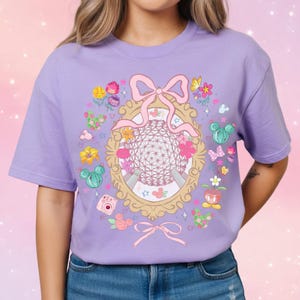 May include: A light purple t-shirt featuring a gold-framed illustration of a geodesic sphere, surrounded by flowers, butterflies, and cacti. A pink bow is at the top and bottom of the frame. The shirt is a short-sleeved crew neck.