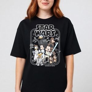 May include: Black t-shirt featuring a Star Wars graphic with Disney characters. The design includes Darth Vader, Mickey Mouse, Donald Duck, and others. The text "STAR WARS" is at the top, with "DESIGNED BY AMYCE" at the bottom.