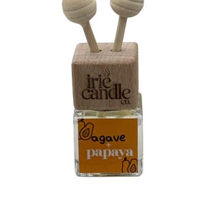 May include: A small square glass bottle containing clear liquid with a wooden cap featuring two wooden spheres on sticks. The label is orange with black text reading agave + papaya and includes line drawings of fruit. The cap is branded irie candle co.