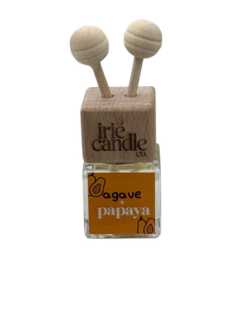 May include: A small square glass bottle containing clear liquid with a wooden cap featuring two wooden spheres on sticks. The label is orange with black text reading agave + papaya and includes line drawings of fruit. The cap is branded irie candle co.