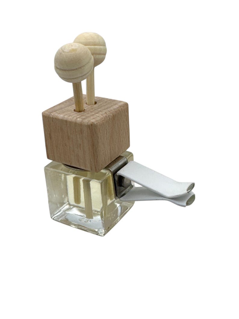 May include: A square glass car air freshener bottle filled with clear liquid, topped with a light-colored wooden block and two small wooden spheres on sticks. A white plastic clip is attached to the side for mounting onto a vehicle vent.