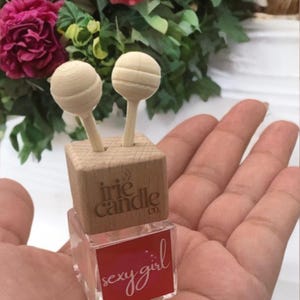May include: A small square glass bottle with a red label reading "sexy girl" and a light wood cap featuring the text "irie candle co." Two light wood spheres on sticks are inserted into the top of the wooden cap, extending upwards from the bottle.