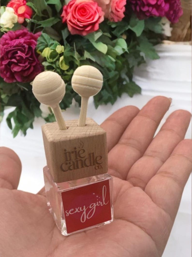 May include: A small square glass bottle with a red label reading "sexy girl" and a light wood cap featuring the text "irie candle co." Two light wood spheres on sticks are inserted into the top of the wooden cap, extending upwards from the bottle.