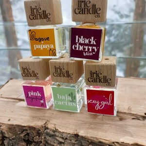 May include: Five square glass car air freshener bottles with wooden caps branded Irie Candle Co. The labels feature various scents and designs: Agave + Papaya, Black Cherry Bliss, Pink Dragonfruit, Baja Cactus, and Sexy Girl with a lip icon.