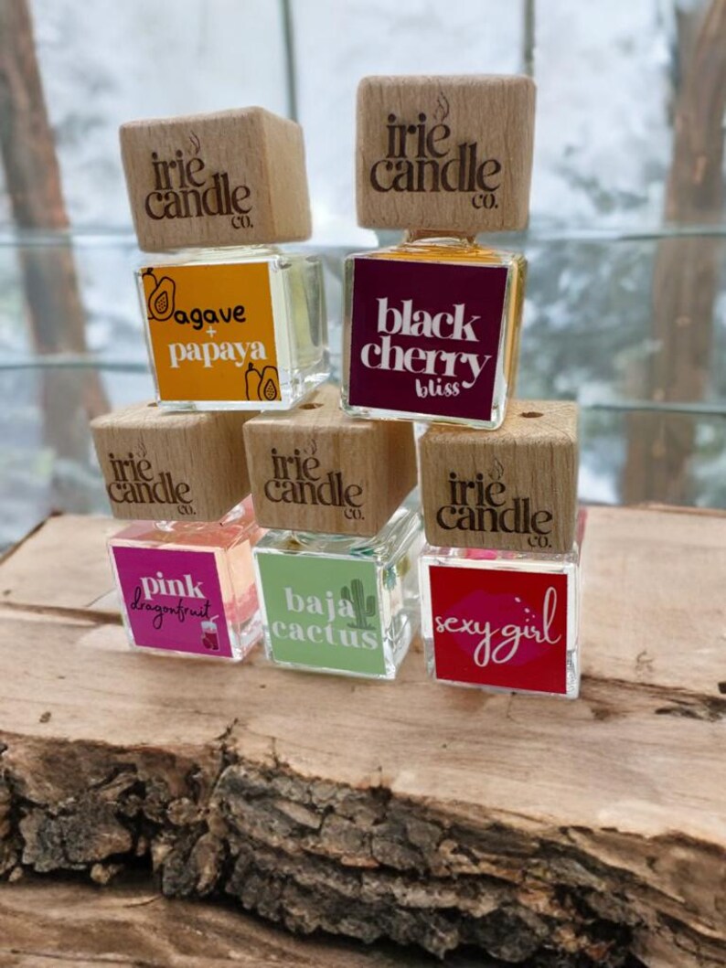 May include: Five square glass car air freshener bottles with wooden caps branded Irie Candle Co. The labels feature various scents and designs: Agave + Papaya, Black Cherry Bliss, Pink Dragonfruit, Baja Cactus, and Sexy Girl with a lip icon.