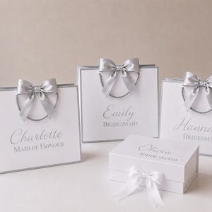 Op de afbeelding: Drie witte cadeauzakjes en één witte cadeaodoos met zilveren rand en zilveren koordhengsels. Elk item is voorzien van een zilveren satijnen strik en gepersonaliseerde tekst: Charlotte Maid of Honour, Emily Bridesmaid, Hannah Bridesmaid en Olivia Bridesmaid.