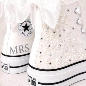 May include: A pair of white high-top sneakers featuring large organza bows at the heels, white satin ribbon laces, and scattered pearl and rhinestone embellishments. The left sneaker displays the text MRS and the Converse All Star circular logo.
