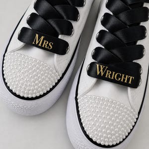 May include: A pair of white canvas sneakers featuring white pearl embellishments on the toe caps, wide black satin ribbon laces, and gold text on the ribbons reading MRS on the left shoe and WRIGHT on the right shoe with silver eyelets.