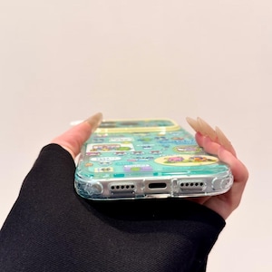 Puede incluir: Funda de tel&eacute;fono transparente con un dise&ntilde;o de dibujos animados colorido. La funda tiene un borde azul claro y presenta varios personajes de dibujos animados. La funda la sostiene una persona con una manga negra.
