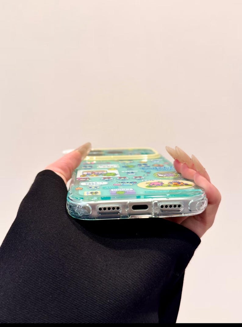 Puede incluir: Funda de tel&eacute;fono transparente con un dise&ntilde;o de dibujos animados colorido. La funda tiene un borde azul claro y presenta varios personajes de dibujos animados. La funda la sostiene una persona con una manga negra.