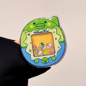 May include: A Tamagotchi virtual pet toy in a green and blue design. The screen displays various cartoon characters. The toy has the text "Tamagotchi" on the front.