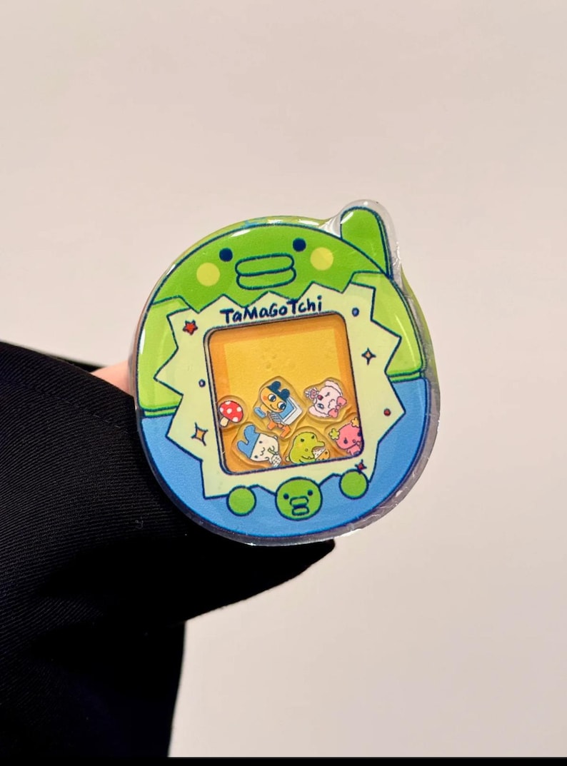 May include: A Tamagotchi virtual pet toy in a green and blue design. The screen displays various cartoon characters. The toy has the text "Tamagotchi" on the front.