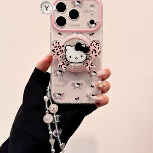 A clear Hello Kitty phone case with a pink bumper is shown front-on with a large Hello Kitty grip and a dangling beaded wristlet.