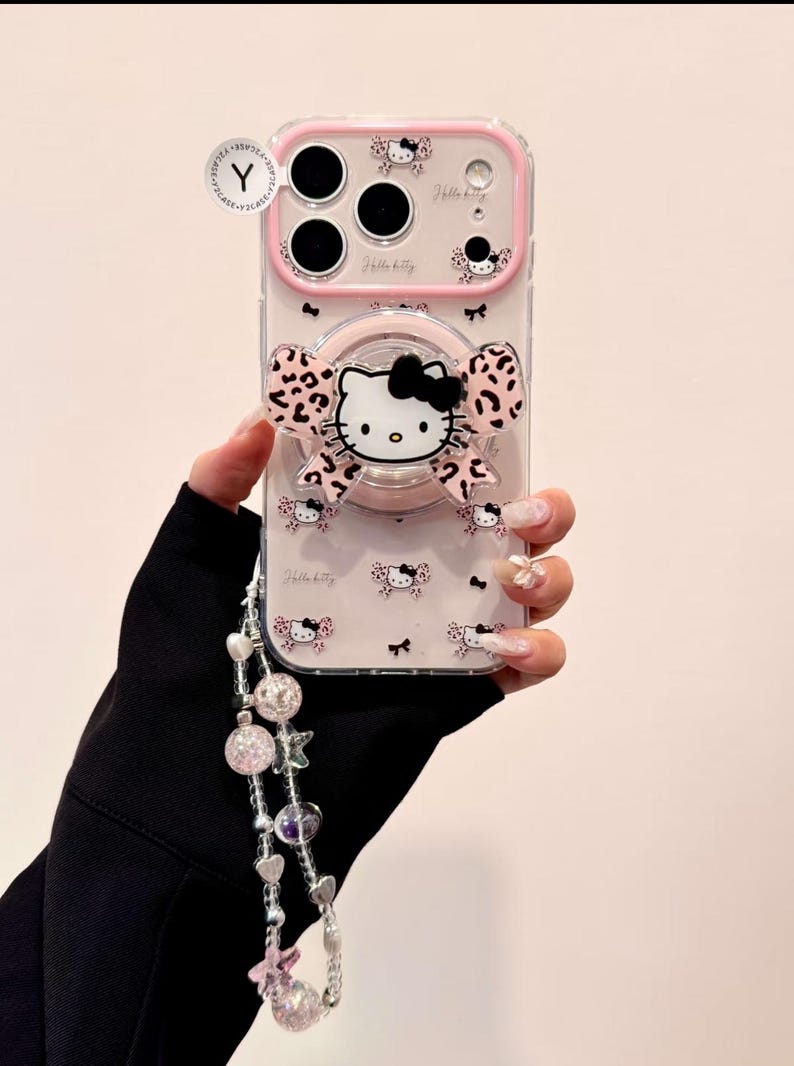 A clear Hello Kitty phone case with a pink bumper is shown front-on with a large Hello Kitty grip and a dangling beaded wristlet.