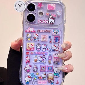May include: Clear phone case with a purple background and Hello Kitty themed decorations. The case features various Hello Kitty images, stars, and other decorative elements. The case has a cat ear design at the top.