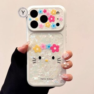 May include: White phone case featuring a Hello Kitty design with a flower crown and colorful floral accents. The case has a pearlescent finish and the text "Hello Kitty" is visible.