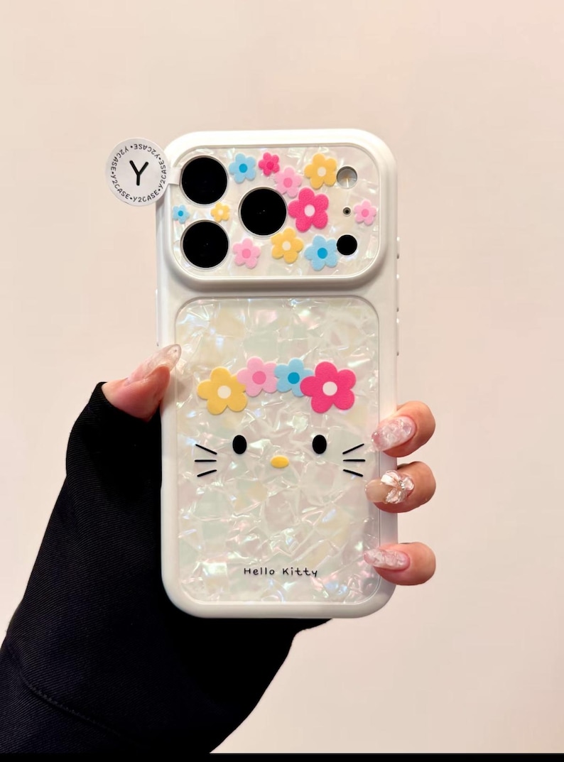 May include: White phone case featuring a Hello Kitty design with a flower crown and colorful floral accents. The case has a pearlescent finish and the text "Hello Kitty" is visible.