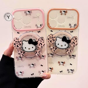 Two clear Hello Kitty phone cases are displayed side by side, one with a pink bumper and one with an orange bumper, each with a large Hello Kitty grip attached.