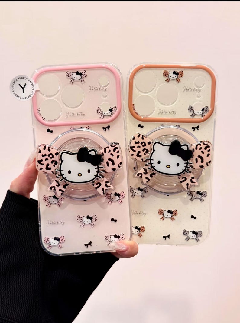 Two clear Hello Kitty phone cases are displayed side by side, one with a pink bumper and one with an orange bumper, each with a large Hello Kitty grip attached.
