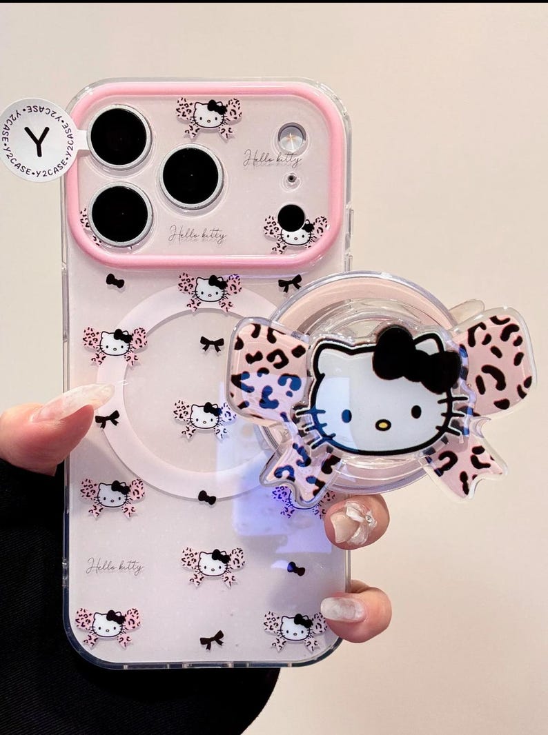 A clear Hello Kitty phone case with a pink bumper is shown held in hand with a large Hello Kitty-shaped grip attached to the back.