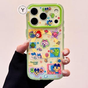 May include: A clear phone case with a green border, adorned with colorful cartoon stickers. The stickers feature various characters, including a red strawberry, a pink bow-wearing character, and a green frog. The text "Tamago Tami" is visible.