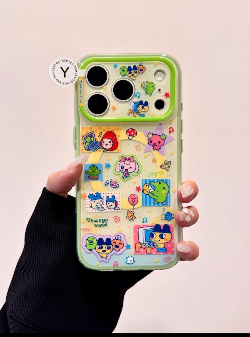 May include: A clear phone case with a green border, adorned with colorful cartoon stickers. The stickers feature various characters, including a red strawberry, a pink bow-wearing character, and a green frog. The text "Tamago Tami" is visible.