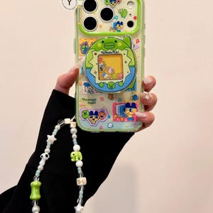 May include: A clear phone case with a green border featuring a Tamagotchi design. The case has a beaded strap with white, green, and brown beads. The Tamagotchi design includes characters and the text "Tamagotchi".
