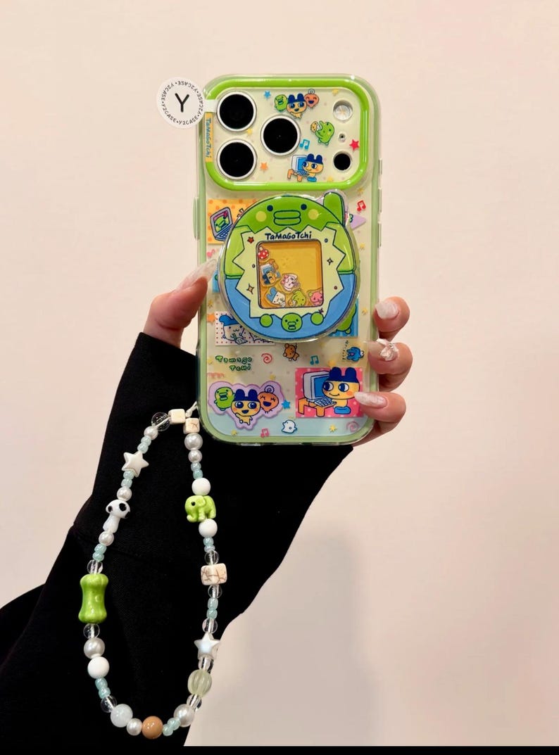 May include: A clear phone case with a green border featuring a Tamagotchi design. The case has a beaded strap with white, green, and brown beads. The Tamagotchi design includes characters and the text "Tamagotchi".