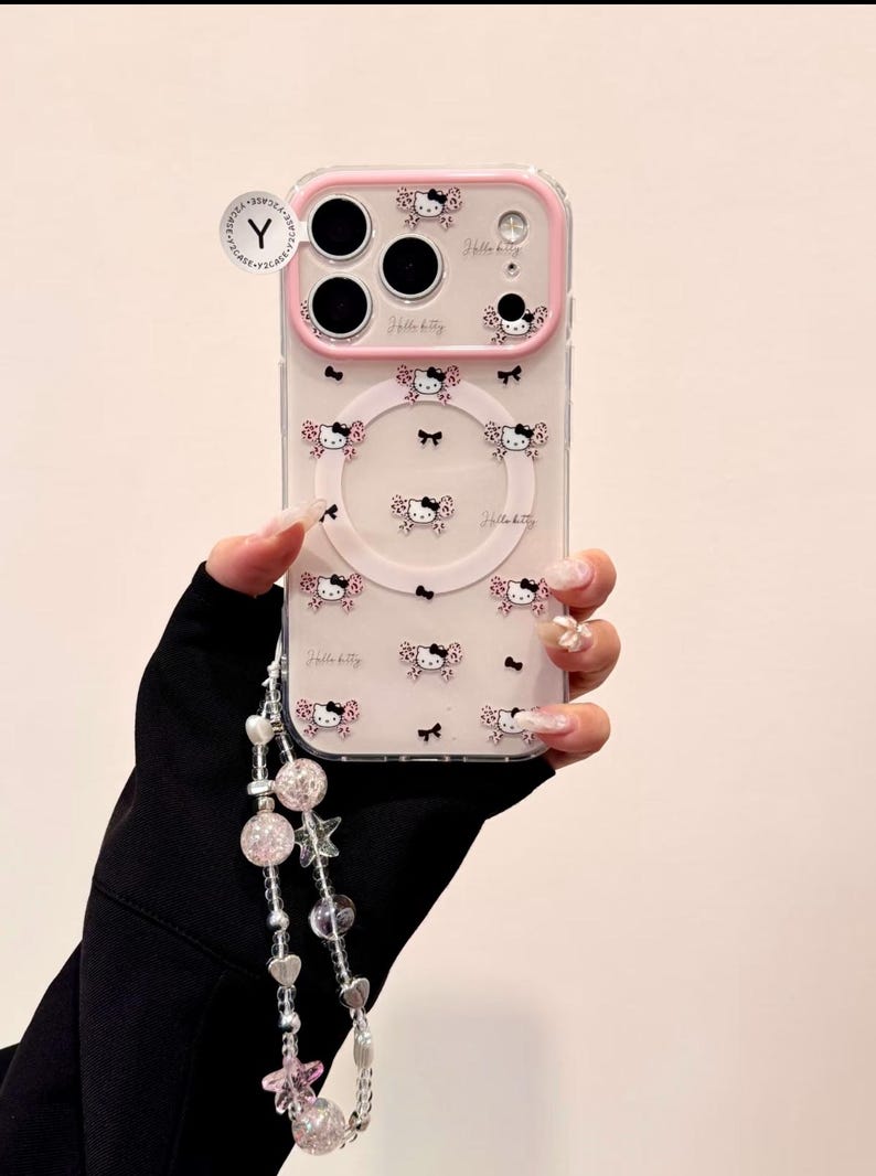 A clear Hello Kitty phone case with a pink bumper is shown front-on with a large Hello Kitty grip and a dangling beaded wristlet.