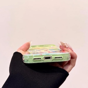 May include: A clear phone case with a colorful cartoon design. The case has a light green border and features a variety of cartoon characters and scenes. The case is held in a hand with long, manicured nails.