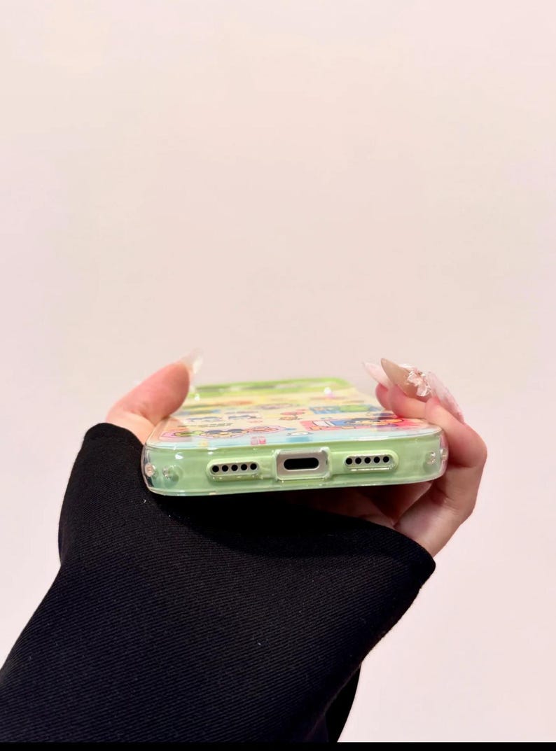 May include: A clear phone case with a colorful cartoon design. The case has a light green border and features a variety of cartoon characters and scenes. The case is held in a hand with long, manicured nails.
