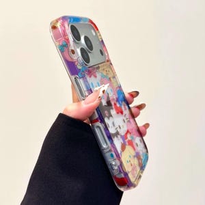 May include: Clear phone case featuring a colorful cartoon character design. The case has a transparent background with a vibrant pattern of various characters in shades of pink, blue, yellow, and purple. The case is designed to fit a smartphone.