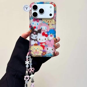 May include: Clear phone case featuring a colorful pattern of cartoon characters. The case has a matching beaded charm with pink and white accents. The phone case is designed to fit a smartphone with three camera lenses.