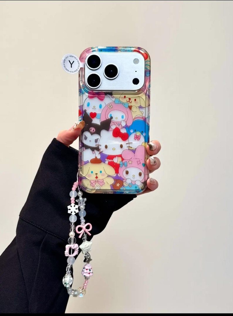 May include: Clear phone case featuring a colorful pattern of cartoon characters. The case has a matching beaded charm with pink and white accents. The phone case is designed to fit a smartphone with three camera lenses.