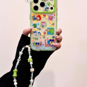 May include: A clear phone case with a green border, decorated with colorful cartoon characters and the text "Tamagotchi". A beaded phone charm with white, green, and brown beads is attached.