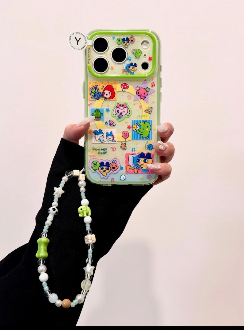 May include: A clear phone case with a green border, decorated with colorful cartoon characters and the text "Tamagotchi". A beaded phone charm with white, green, and brown beads is attached.